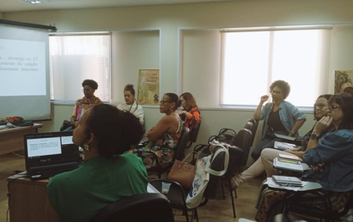 #DescriçãoDaImagem Na imagem, aparecem conselheiras do CRESS-RJ assistindo à apresentação da Seccional Norte Fluminense. A conselheira Ully Azevedo, da Seccional, uma mulher parda vestindo blusa verde, está sentada com um laptop em uma bancada a sua frente fazendo a apresentação. As outras conselheiras olham par Ully e assistem à apresentação. Há cadeiras azuis espalhadas no auditório e as paredes são claras.